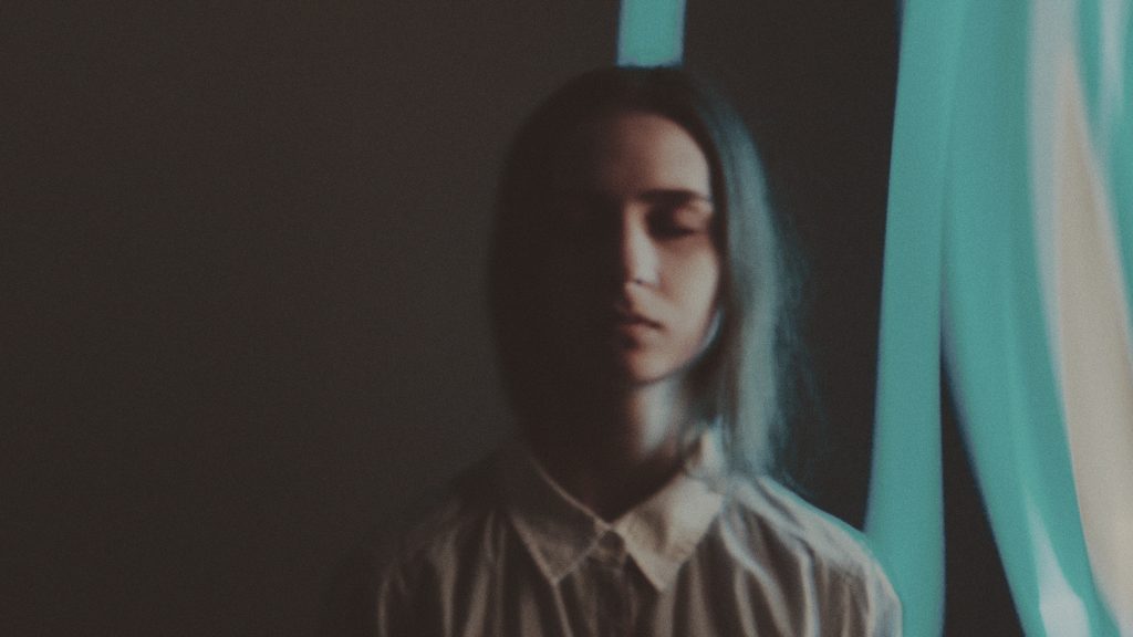 A person with long hair stands in soft light, eyes closed, against a dark background with blue ribbons.