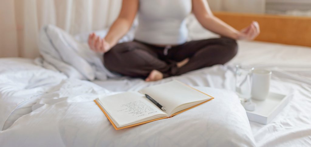 A person meditates on a bed, with a journal and pen nearby, and a cup on a tray beside them.