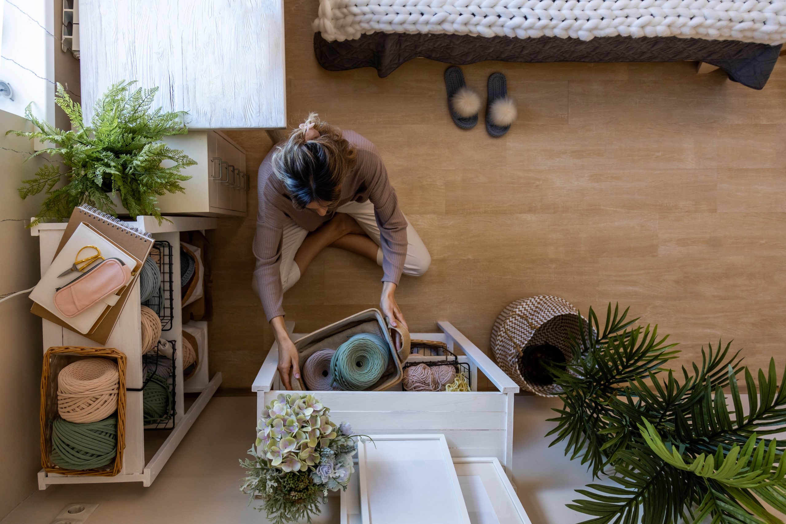 A person organizes a drawer filled with colorful yarn in a cozy, well-decorated room with plants and a bed.