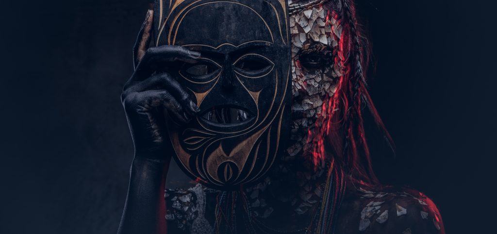 A person partially obscured by a decorative mask, with intricate body paint and dark, textured skin.