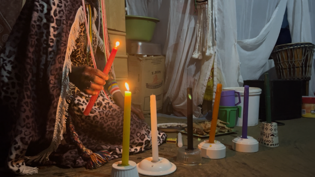 A person in a patterned shawl holds a red candle, surrounded by colorful candles on a table in a dimly lit room.