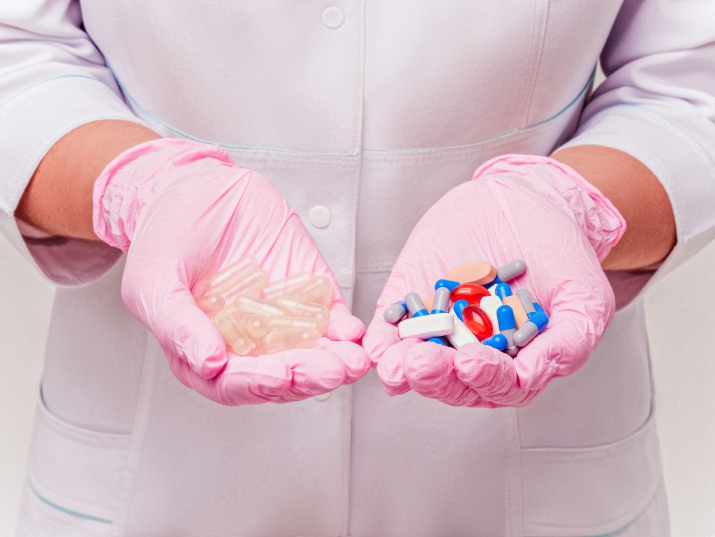 A person in pink gloves holds two handfuls of various pills and capsules, showcasing different colors and shapes.