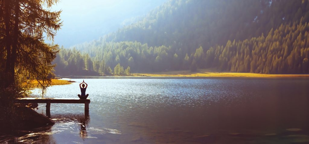 A person practices yoga on a dock by a serene lake, surrounded by lush mountains and soft morning light.