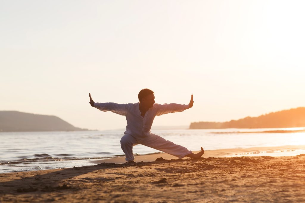 A person practicing martial arts on a beach at sunset, with water and hills in the background.