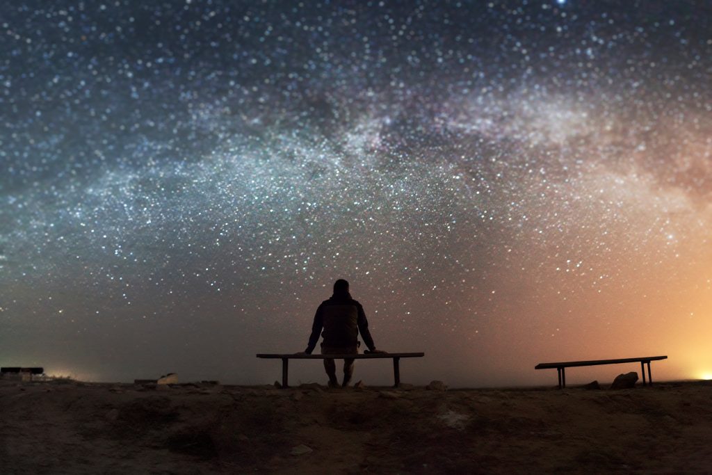 A person sits on a bench, gazing at a star-filled sky with a vibrant Milky Way and distant lights on the horizon.