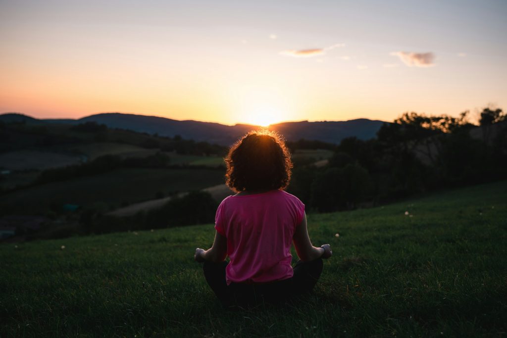A person sits cross-legged on grass, watching a sunset over rolling hills, with a serene expression and curly hair.