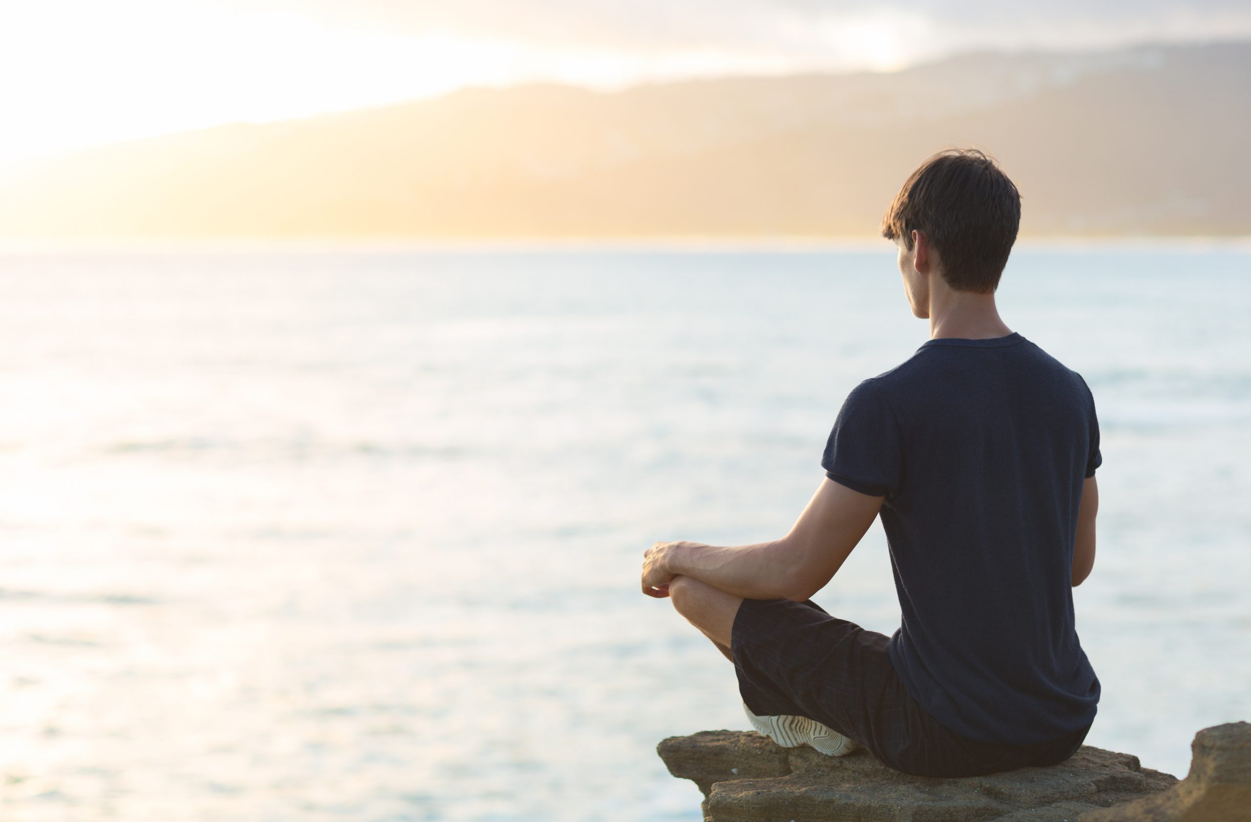A person sits cross-legged on a rock by the water, gazing at a serene sunset over the horizon.