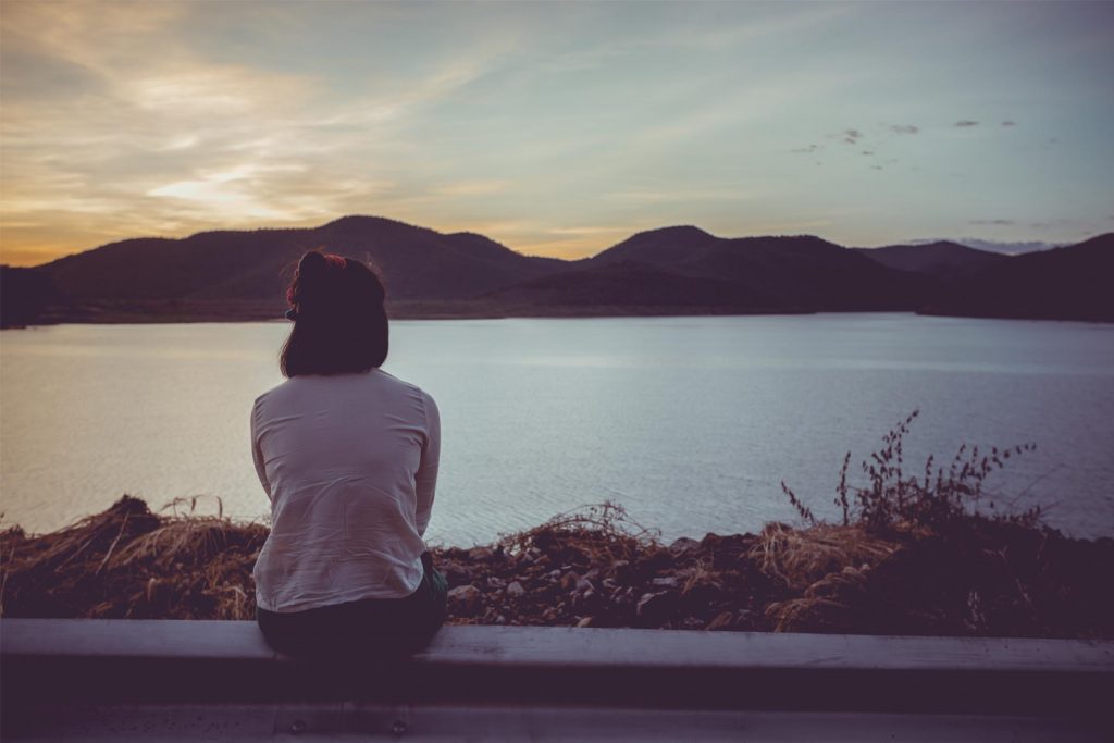 A person sits by a lake at sunset, gazing at the tranquil water and distant mountains.