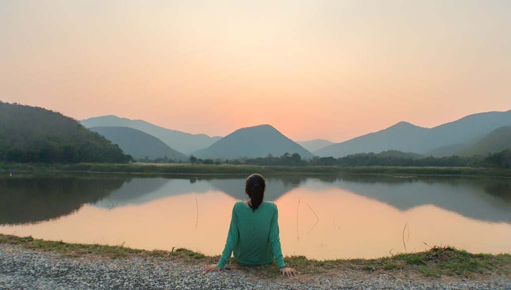 A person sits by a tranquil lake, gazing at a sunset behind distant mountains, reflecting on the water's surface.