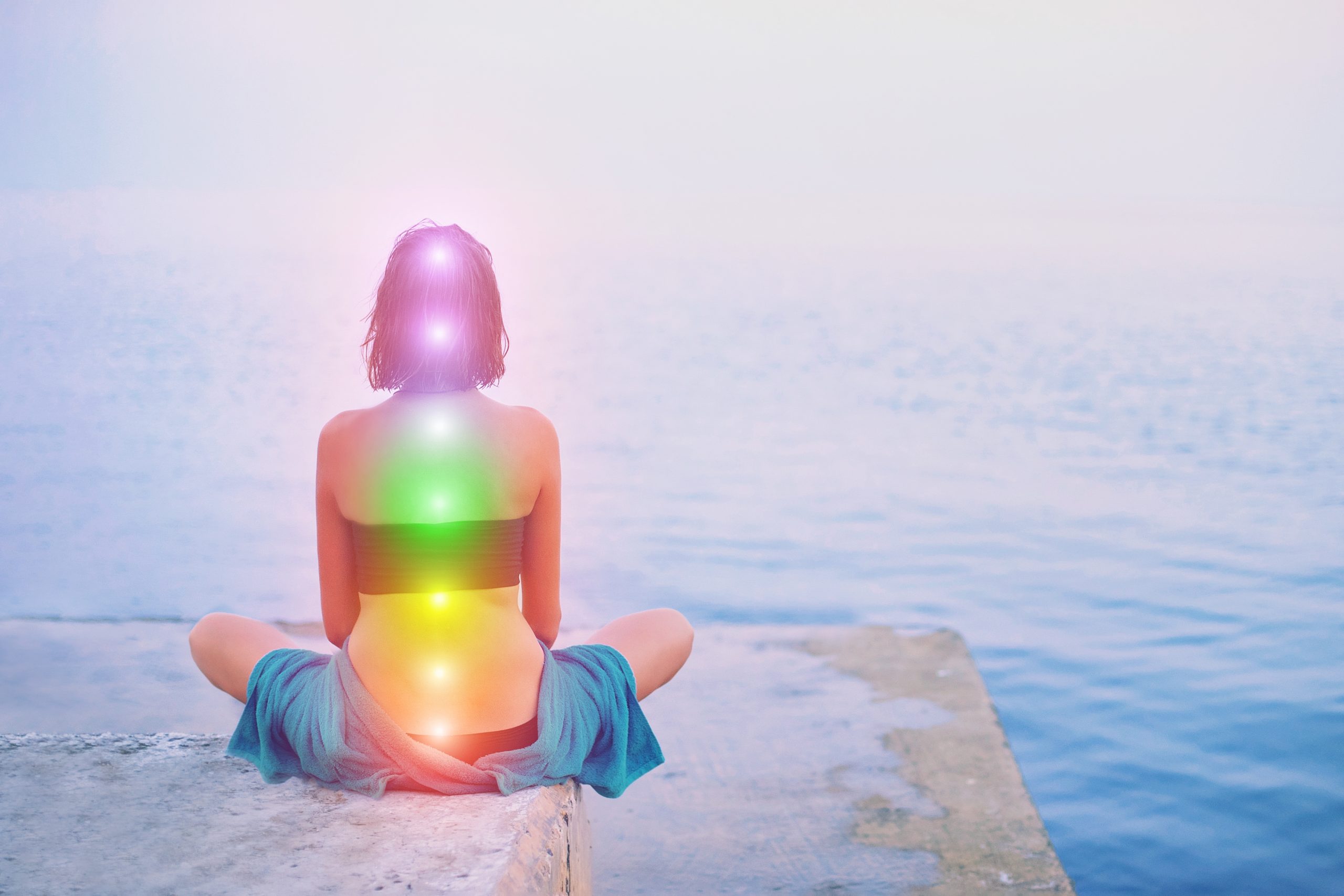 A person sits by the water, facing away, with colorful chakra lights glowing along their spine against a misty backdrop.