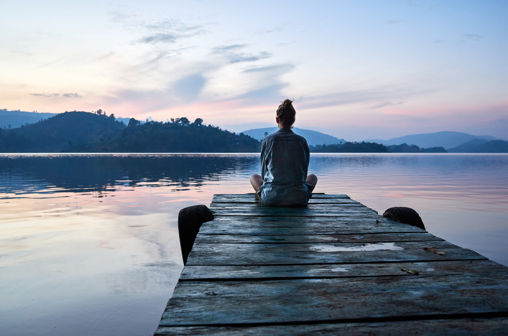 A person sits on a wooden dock, gazing at a serene lake and pastel sky at dawn, surrounded by rolling hills.