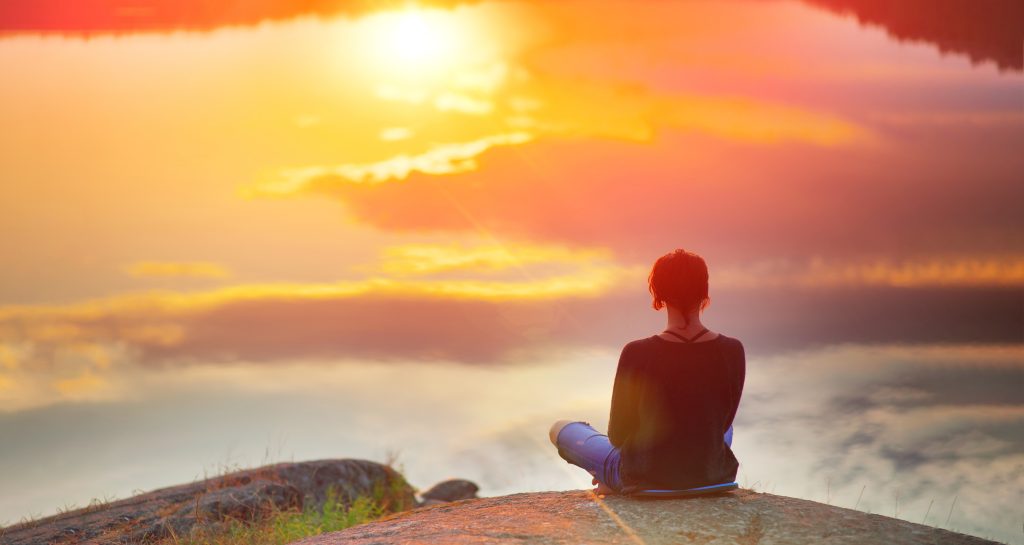 Person sitting on a rock by a lake, gazing at a vibrant sunset reflecting on the water.
