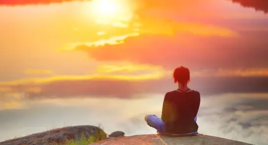 Person sitting on a rock by a serene lake, gazing at a vibrant sunset reflecting on the water.