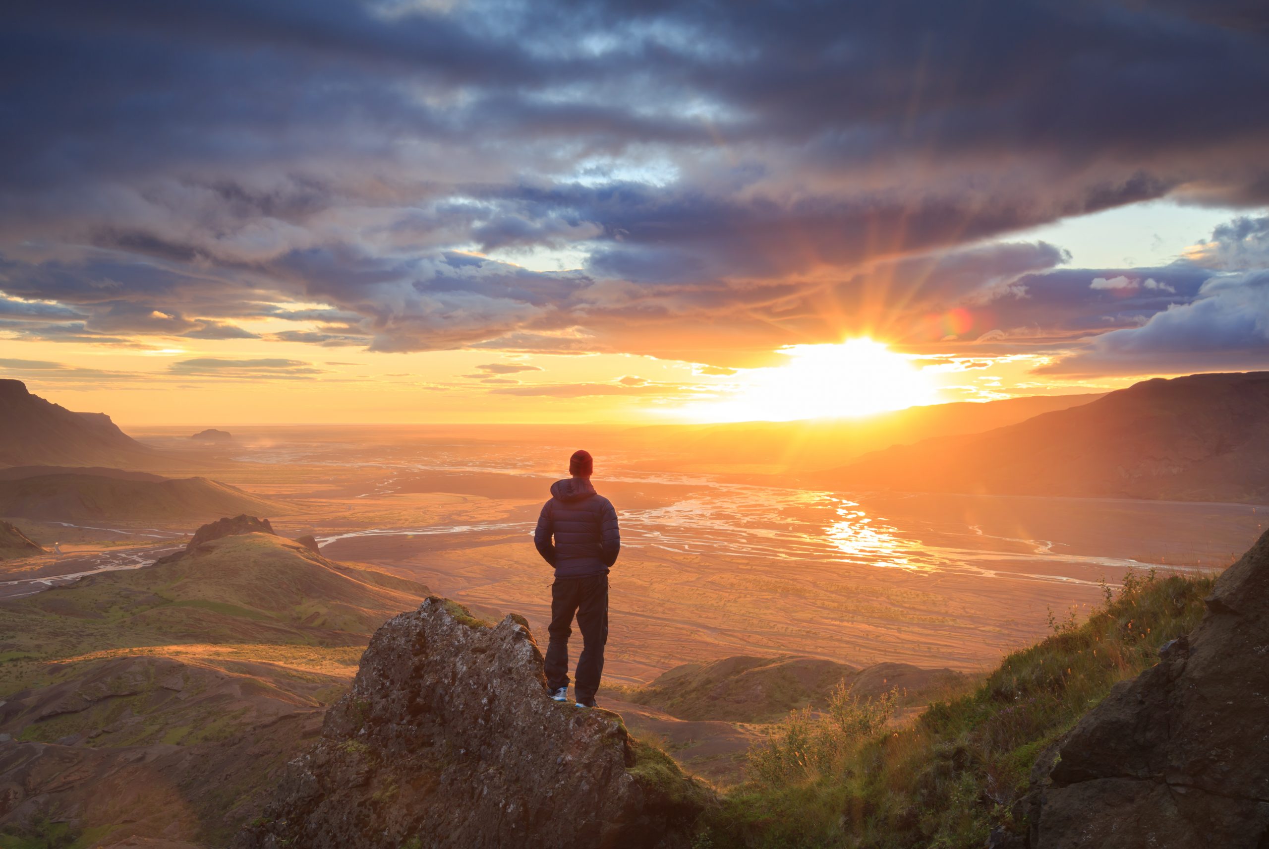 A person stands on a rocky outcrop, gazing at a vibrant sunset over a valley, with dramatic clouds and golden light.