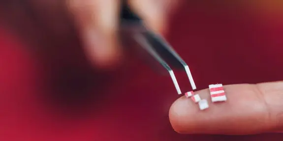 A person using tweezers to hold tiny red and white striped pieces on their fingertip against a blurred background.
