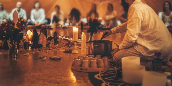 A person in white prepares a potion in a cozy, candlelit space surrounded by flowers and participants seated in the backgr...