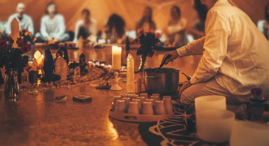 A person in white prepares a potion in a cozy, candlelit space surrounded by flowers and participants seated in the backgr...