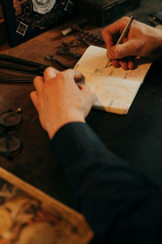 A person writes in a planner on a wooden desk surrounded by vintage tools and glasses.