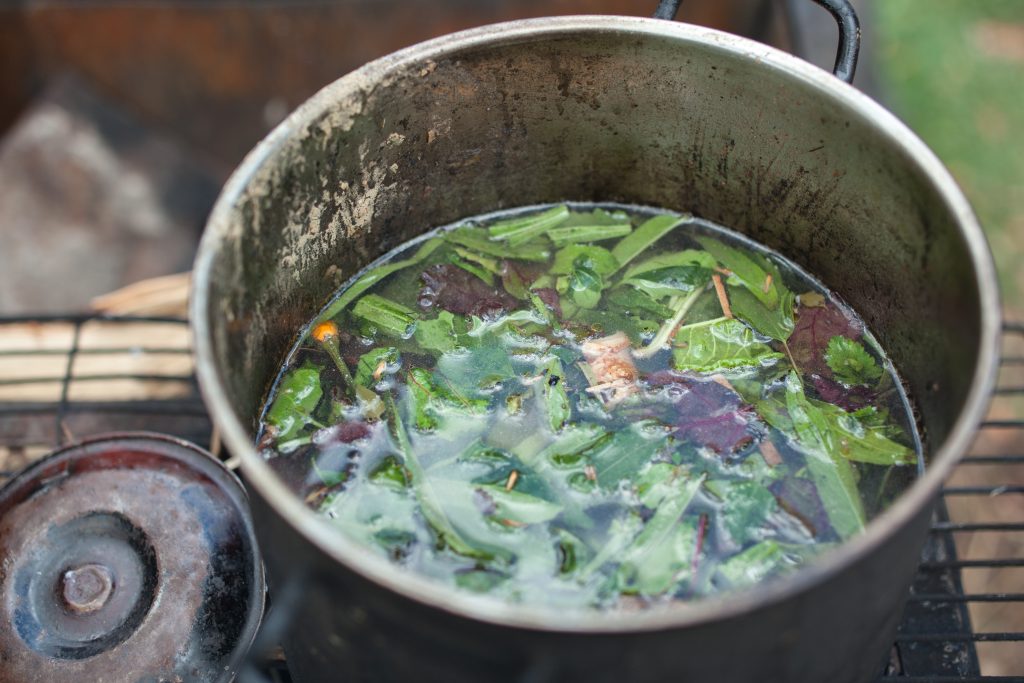 A pot simmering with green herbs and leafy vegetables in a rustic outdoor setting.