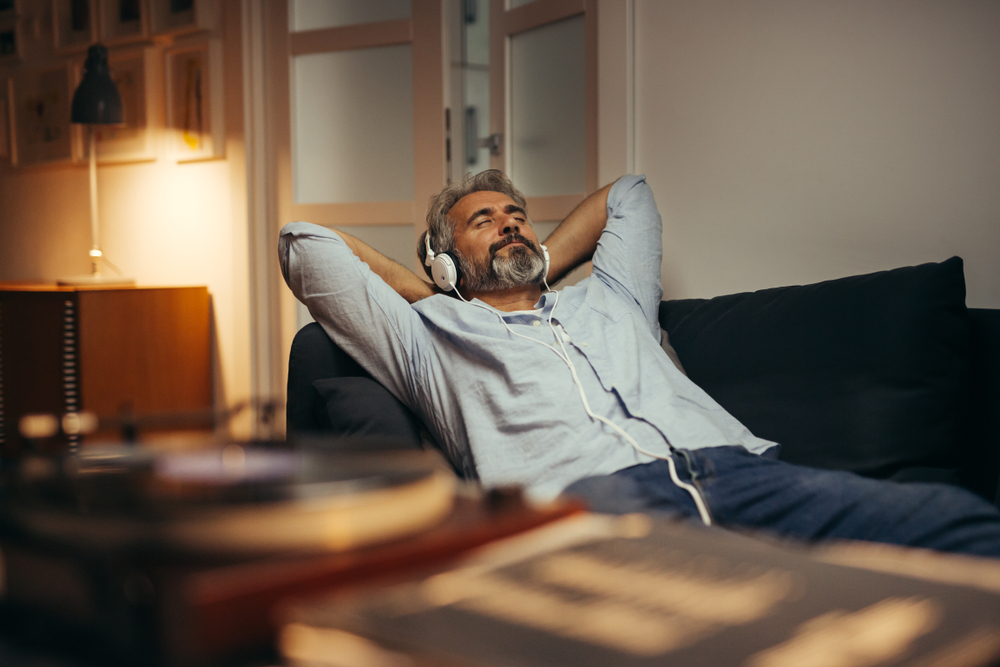 Relaxed man with gray hair and beard listens to music on headphones while lounging on a couch in a cozy room.