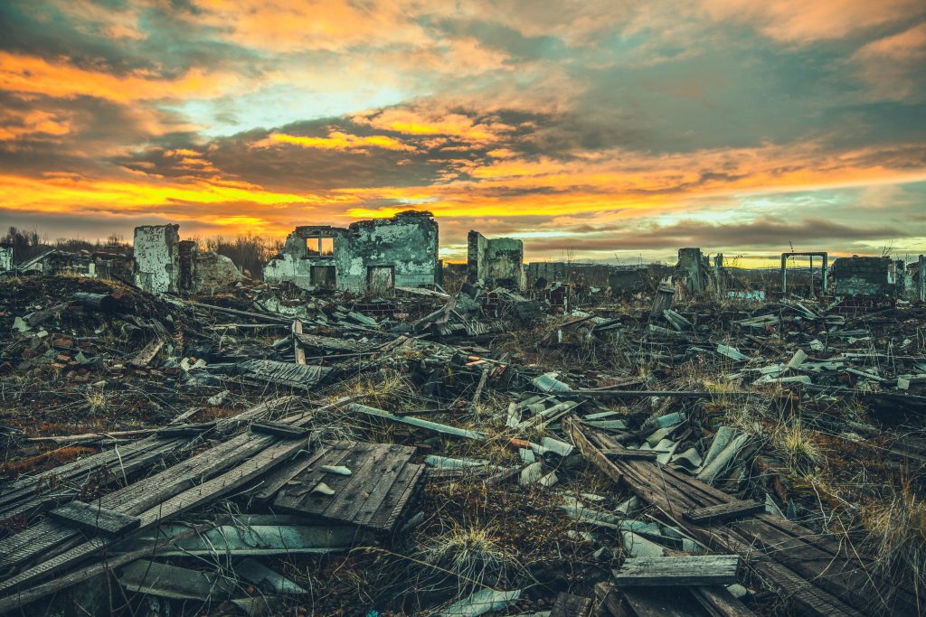 Ruins of a dilapidated building amidst scattered debris, under a dramatic sunset sky with vibrant orange and purple hues.