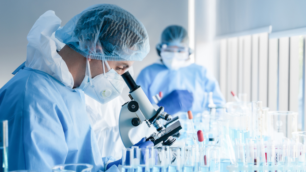 A scientist in protective gear examines samples under a microscope, surrounded by glassware filled with colorful liquids.