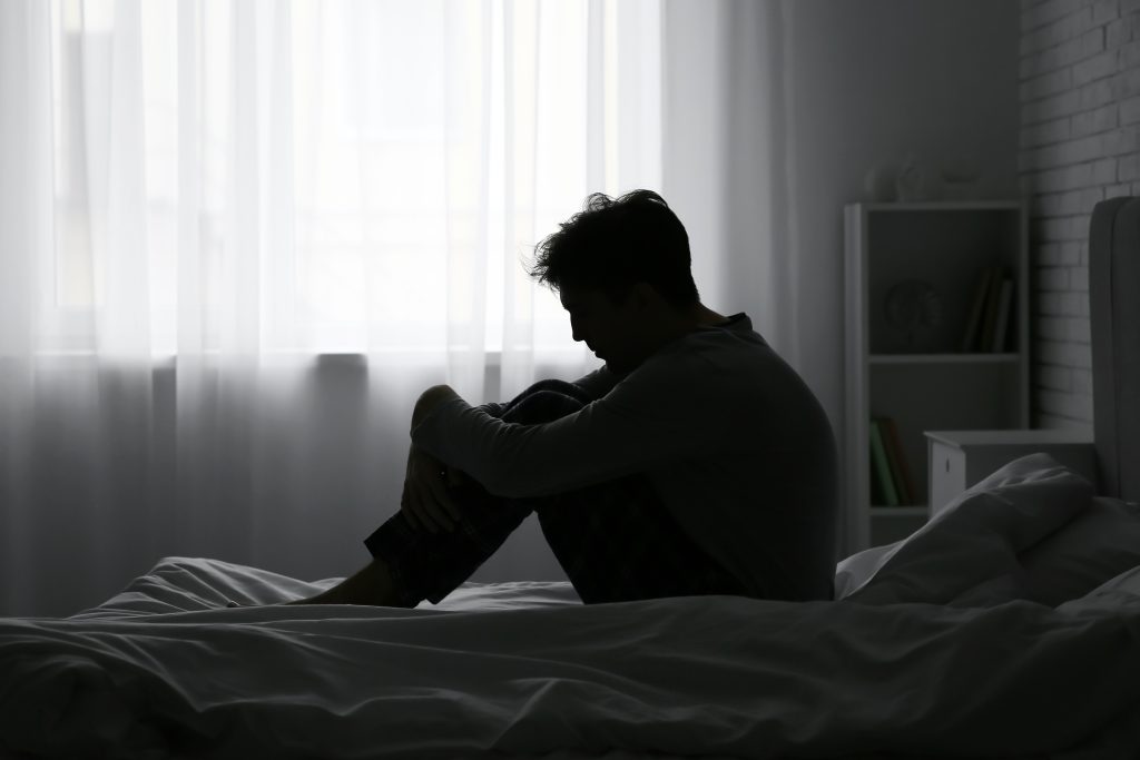 Silhouette of a man sitting on a bed, hugging his knees, with soft light filtering through sheer curtains.