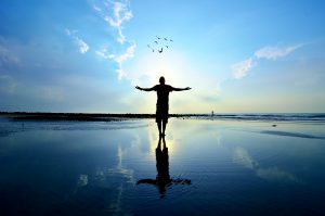 Silhouette of a person with arms outstretched against a sunset, reflecting on calm water with birds flying above.