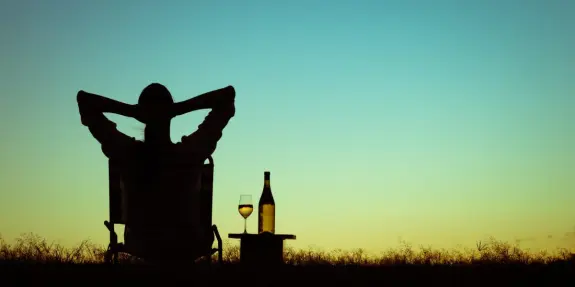 Silhouette of a person relaxing in a chair, with a glass of wine and a bottle on a table against a sunset sky.