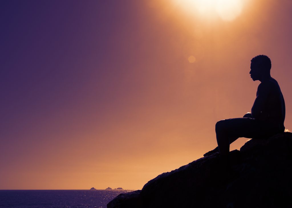 Silhouette of a person sitting on a rock, gazing at the ocean under a vibrant sunset sky.