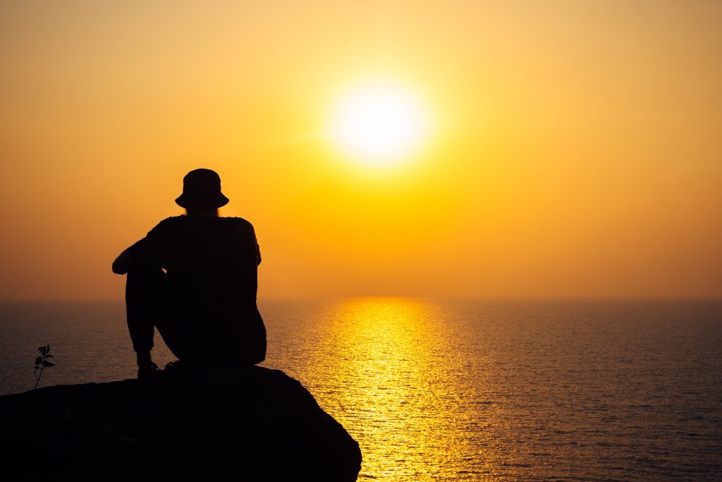 Silhouette of a person sitting on a rock, gazing at a vibrant sunset over the calm ocean.