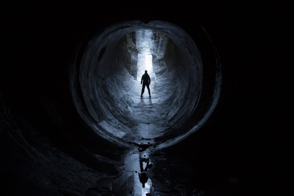 Silhouette of a person standing in a dark tunnel, illuminated by light at the far end, reflecting on wet ground.