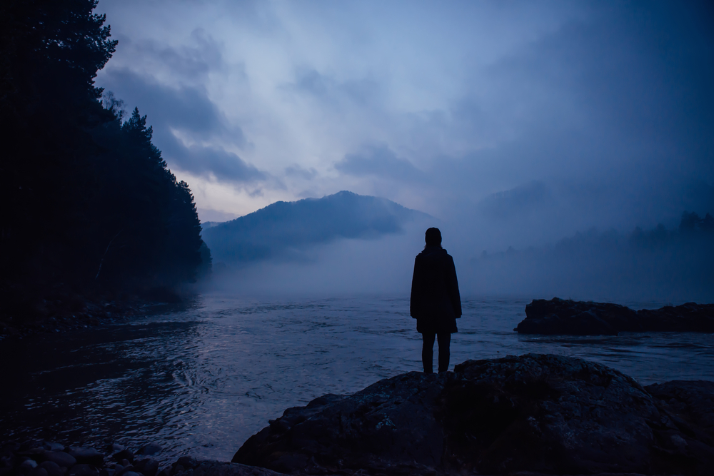 Silhouette of a person standing on a rock by a misty river at twilight, surrounded by mountains and trees.