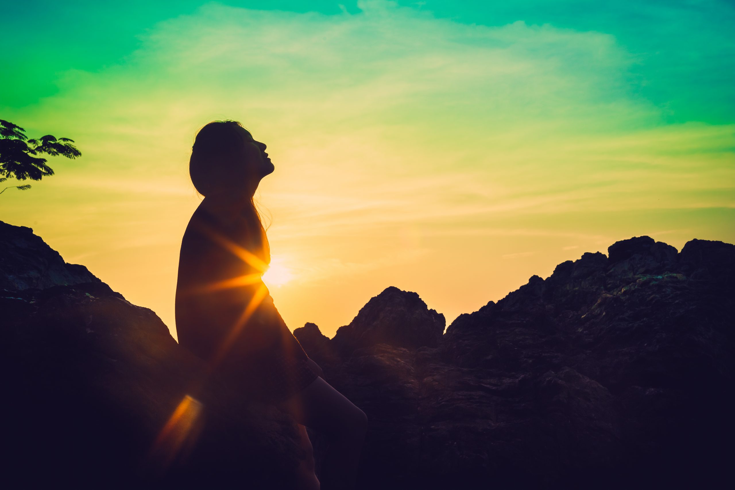 Silhouette of a woman against a vibrant sunset, with sunlight glinting off rocks and a colorful sky.