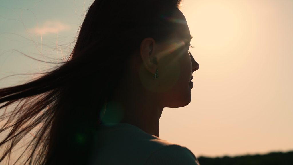 Silhouette of a woman with flowing hair against a bright sunset, capturing a serene and contemplative moment.