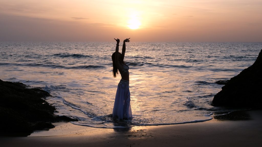 Silhouette of a woman in a flowing white dress, arms raised, against a sunset over the ocean.