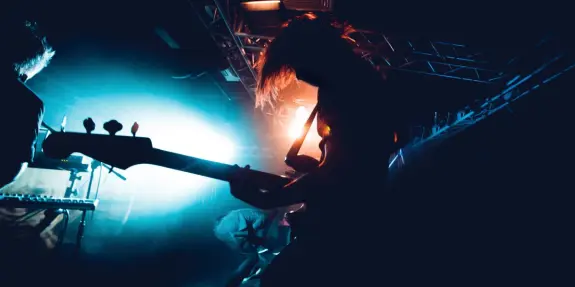 Silhouetted musician playing guitar on stage, illuminated by vibrant blue and orange lights. Energetic atmosphere.