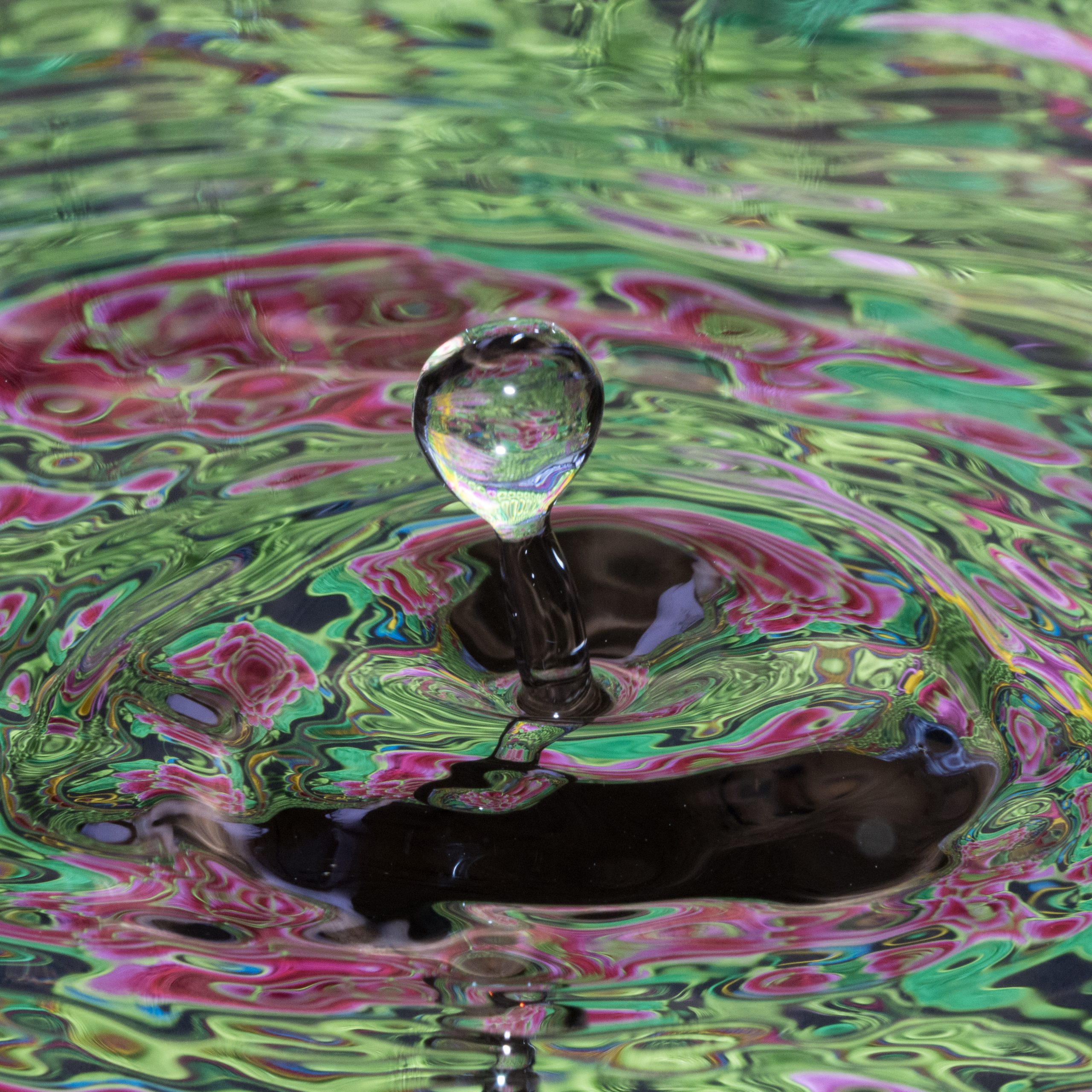 A single water droplet creates ripples on a colorful, swirling surface of pink and green hues.