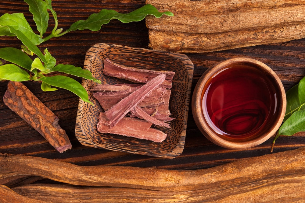 Sliced herbal bark and a small bowl of reddish liquid on a wooden surface, surrounded by green leaves.
