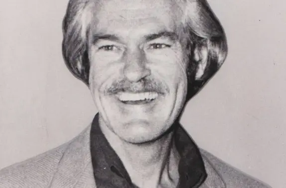 Smiling man with gray hair and a mustache, wearing a light-colored blazer over a dark shirt.