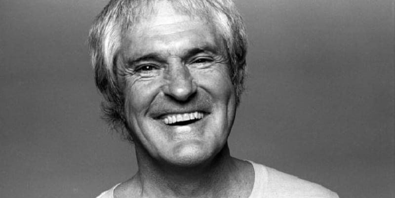 Smiling older man with gray hair, wearing a light shirt, against a plain background.