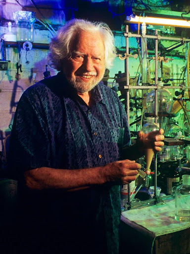 Smiling scientist in a lab, holding a flask, surrounded by glassware and colorful lighting.