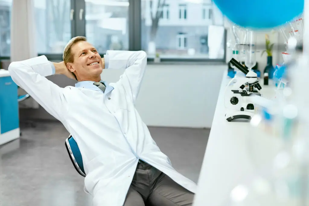 A smiling scientist relaxes in a lab, surrounded by microscopes and glassware, with large windows in the background.