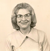 Smiling woman with glasses and wavy hair, wearing a light-colored collared blouse.