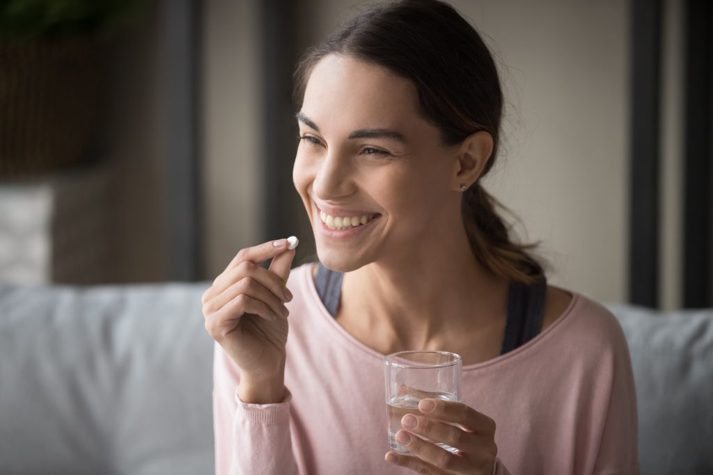 Smiling woman holding a pill and a glass of water, seated in a cozy indoor setting.