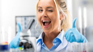 Smiling woman in a lab coat winks and gives thumbs up, surrounded by lab equipment and a microscope.