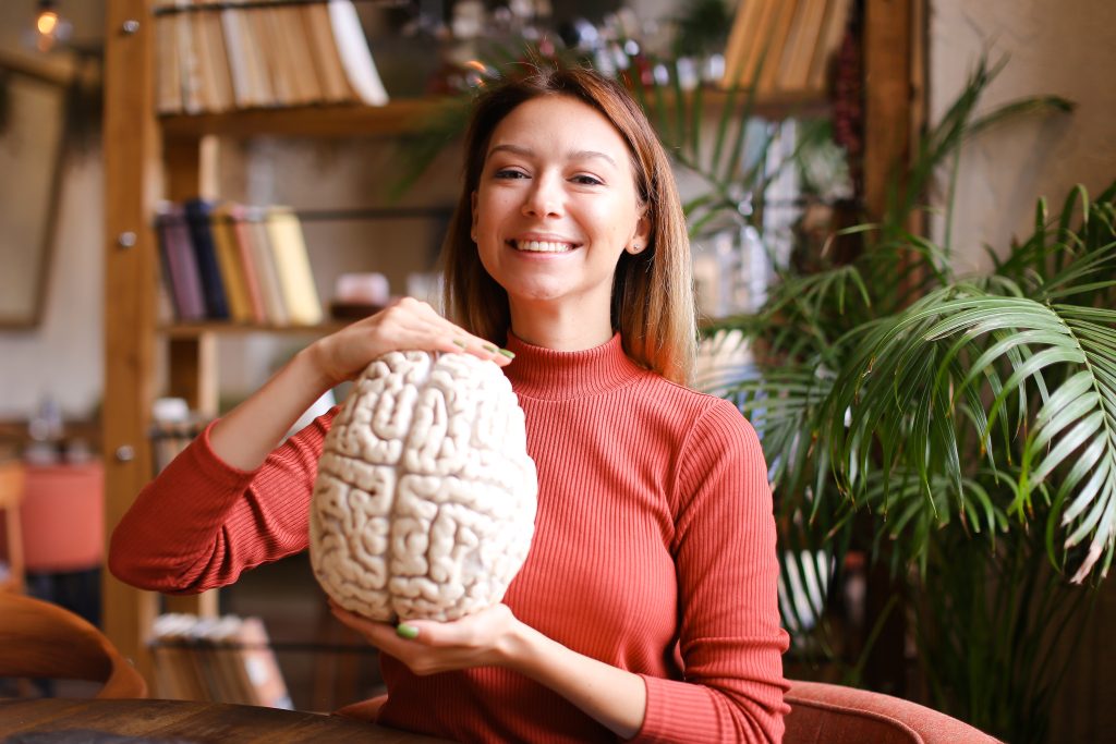 Smiling woman in a red sweater holds a brain-shaped sculpture in a cozy caf&eacute; surrounded by books and plants.