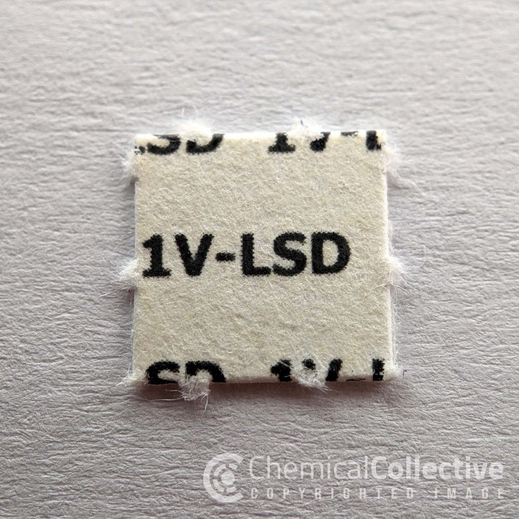 Square blotter paper with printed text "1V-LSD" on a light background. Edges appear slightly frayed.