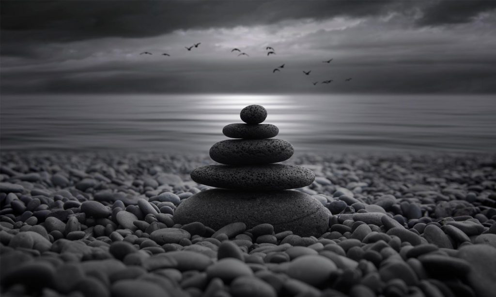 Stacked black stones on a pebbled shore, with a calm sea and birds flying against a moody, cloudy sky.