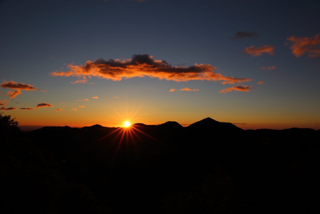 Sunset over mountain silhouettes, with vibrant orange and purple hues illuminating the sky and clouds.
